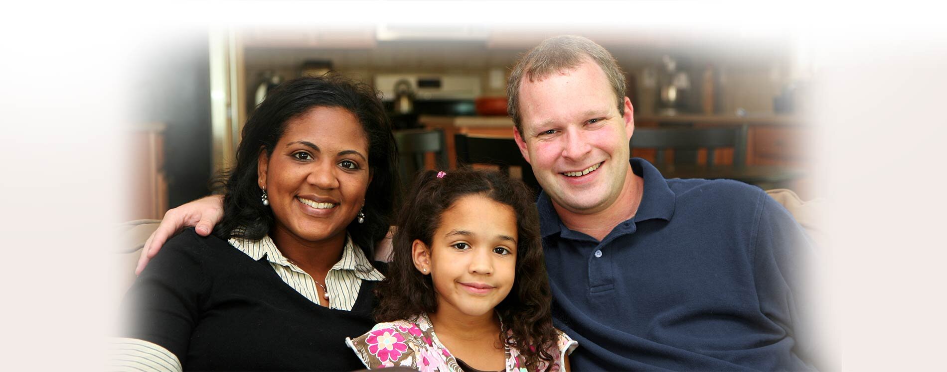 A husband and wife couple sitting on a couch with their daughter between the two.