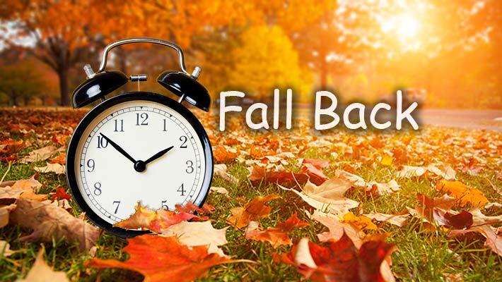 An alarm clock in the grass with fall leaves and the words Fall Back.